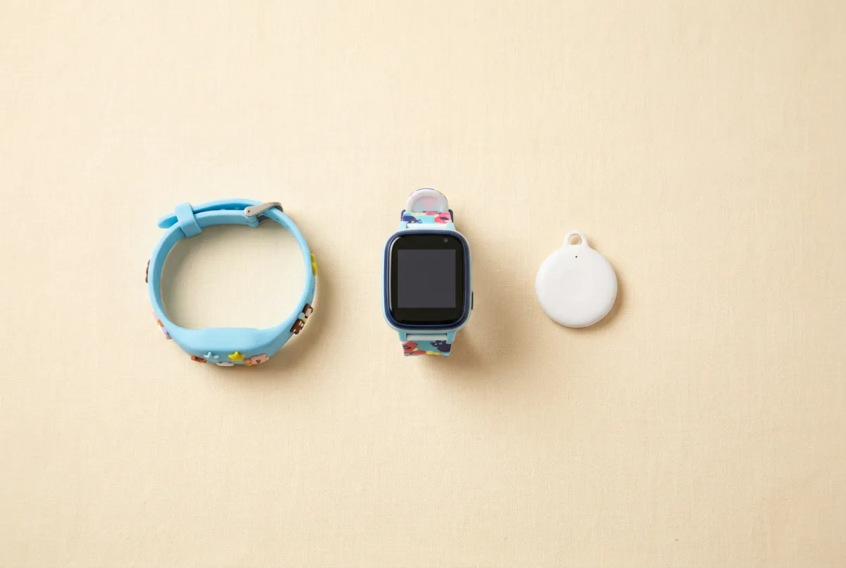 Flat-lay comparison of a kids GPS bracelet, a smartwatch and a small round Bluetooth tracker disc