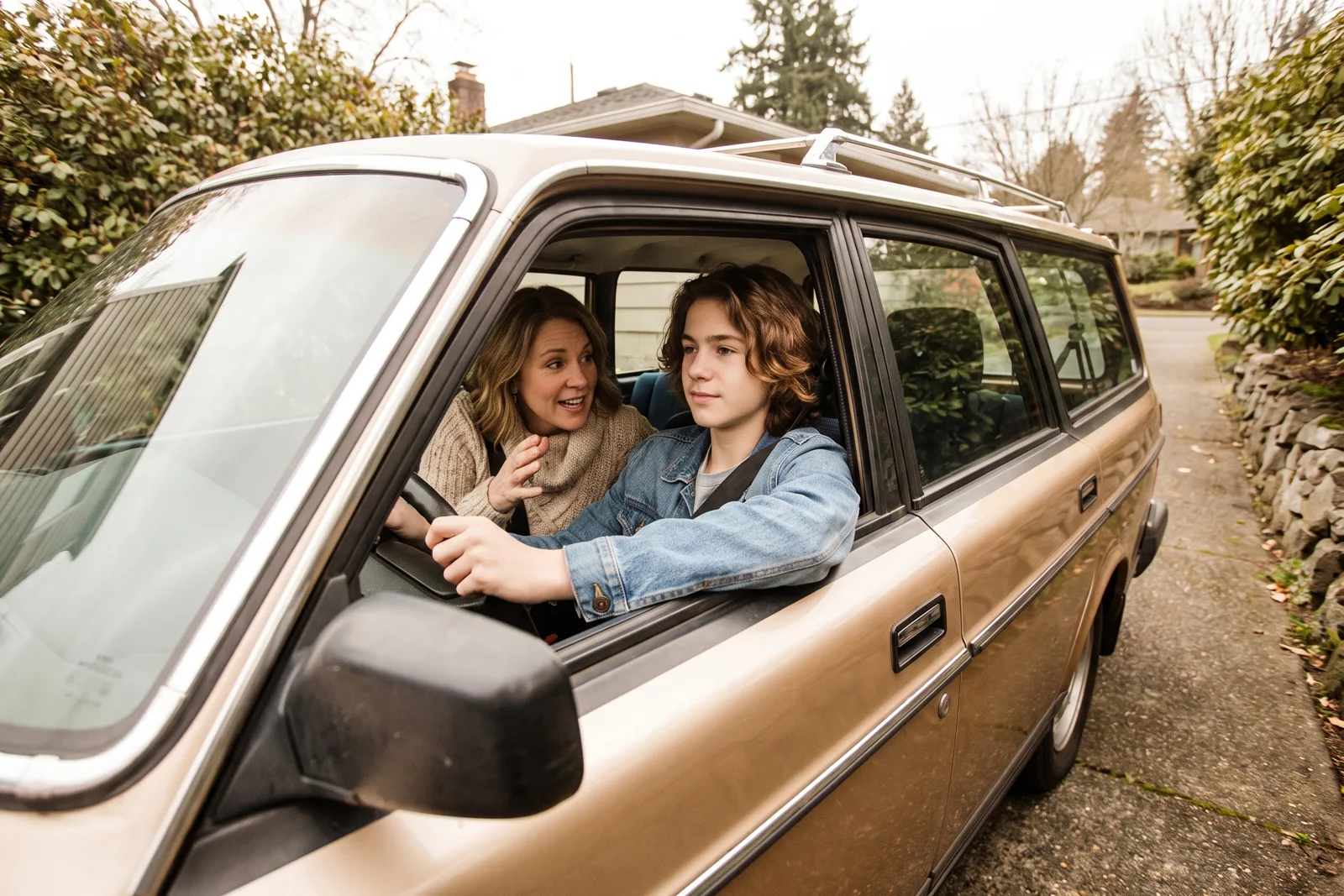 A teenage driver at the wheel of a parked car in a driveway while a parent in the passenger seat talks with them